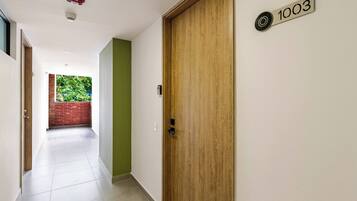 Ein Flur mit einer Holztür mit der Aufschrift '1003', einer grünen Wand und Blick auf eine Ziegelwand und Grünflächen im Freien.
