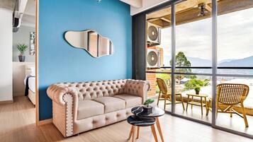 Una sala de estar moderna con un sofá color beige, una mesa redonda con una planta en maceta, y vistas de la ciudad a través de grandes ventanales.