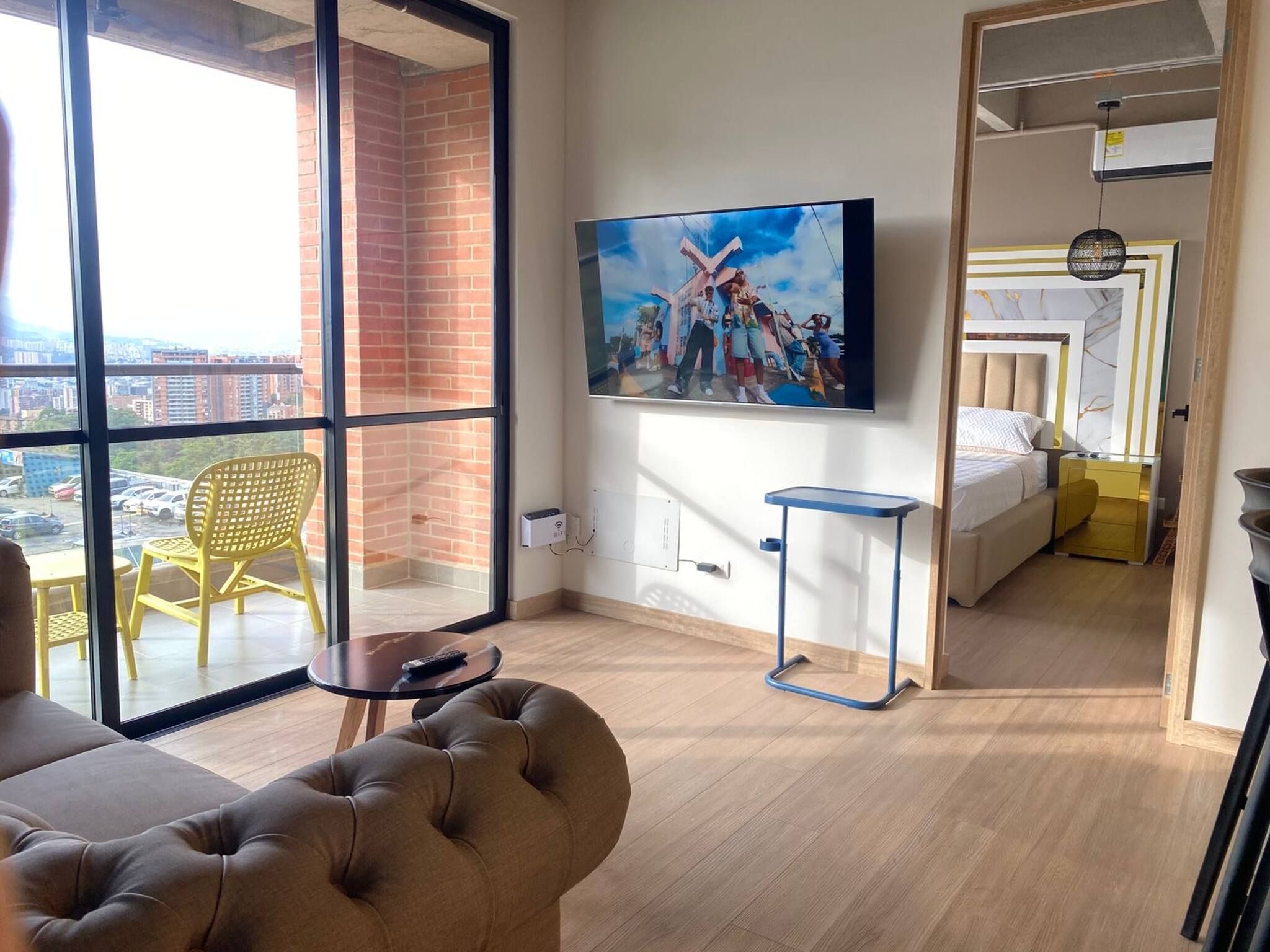 Una habitación de hotel moderna con un ventanal grande, un televisor de pantalla plana, un sofá, una mesita pequeña y una cama con cabecera.