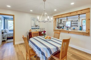 A dining area with a striped tablecloth, wooden chairs, a chandelier, and a kitchen visible through a large window. - Lakefront Adirondack Escape! Fire Pit & Game Room (Broadalbin)