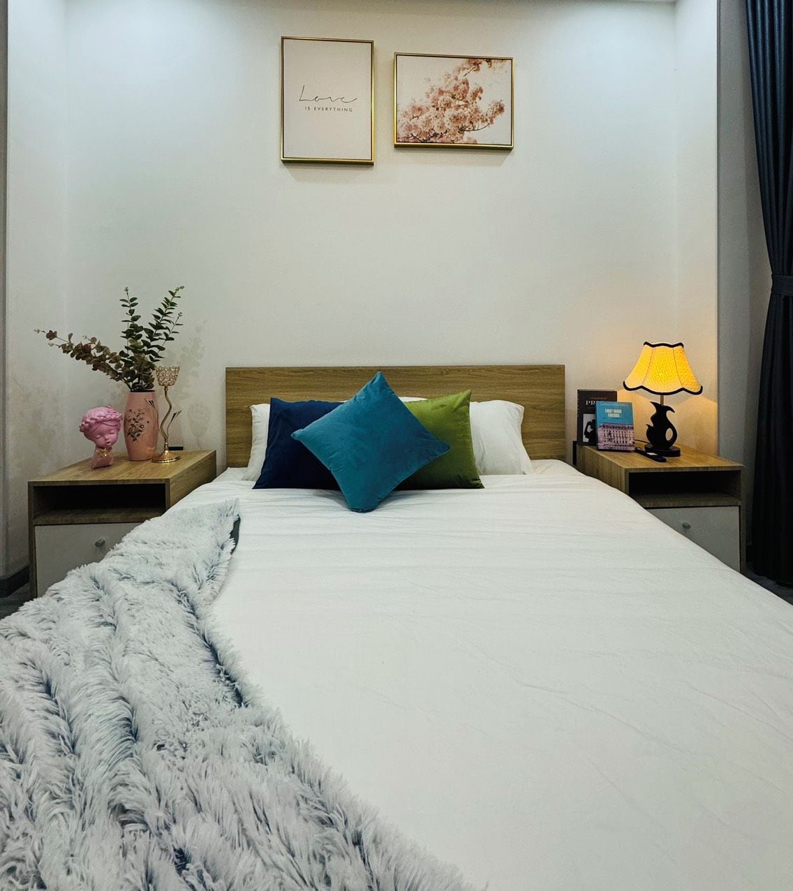 Un lit bien fait, agrémenté d’un jeté blanc moelleux, d’une tête de lit en bois et de coussins colorés. On peut voir des photos encadrées au mur, un vase rempli de fleurs et une lampe sur la table de chevet.