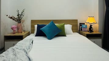 Un lit bien fait, agrémenté d’un jeté blanc moelleux, d’une tête de lit en bois et de coussins colorés. On peut voir des photos encadrées au mur, un vase rempli de fleurs et une lampe sur la table de chevet.