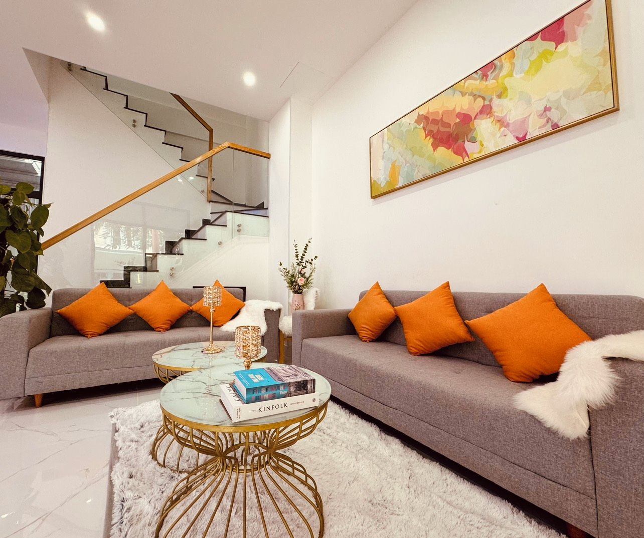 Un salon moderne avec un canapé gris, des coussins orange, une table basse en verre et un escalier en arrière-plan.