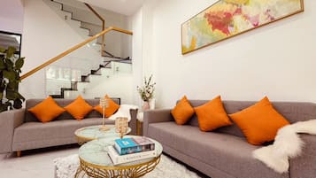 Un salon moderne avec un canapé gris, des coussins orange, une table basse en verre et un escalier en arrière-plan.