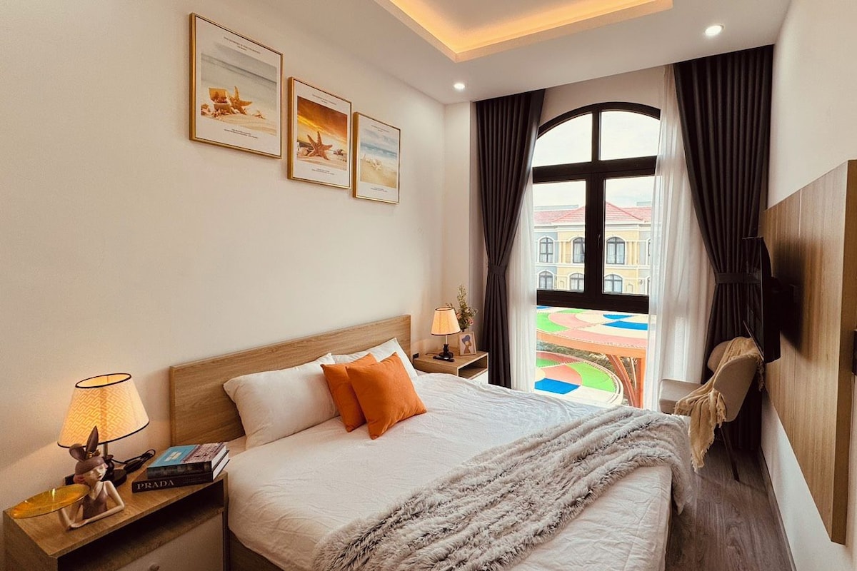 Quarto de hotel com uma cama grande, mesa de cabeceira com luminária, estante de livros e vista para o exterior.