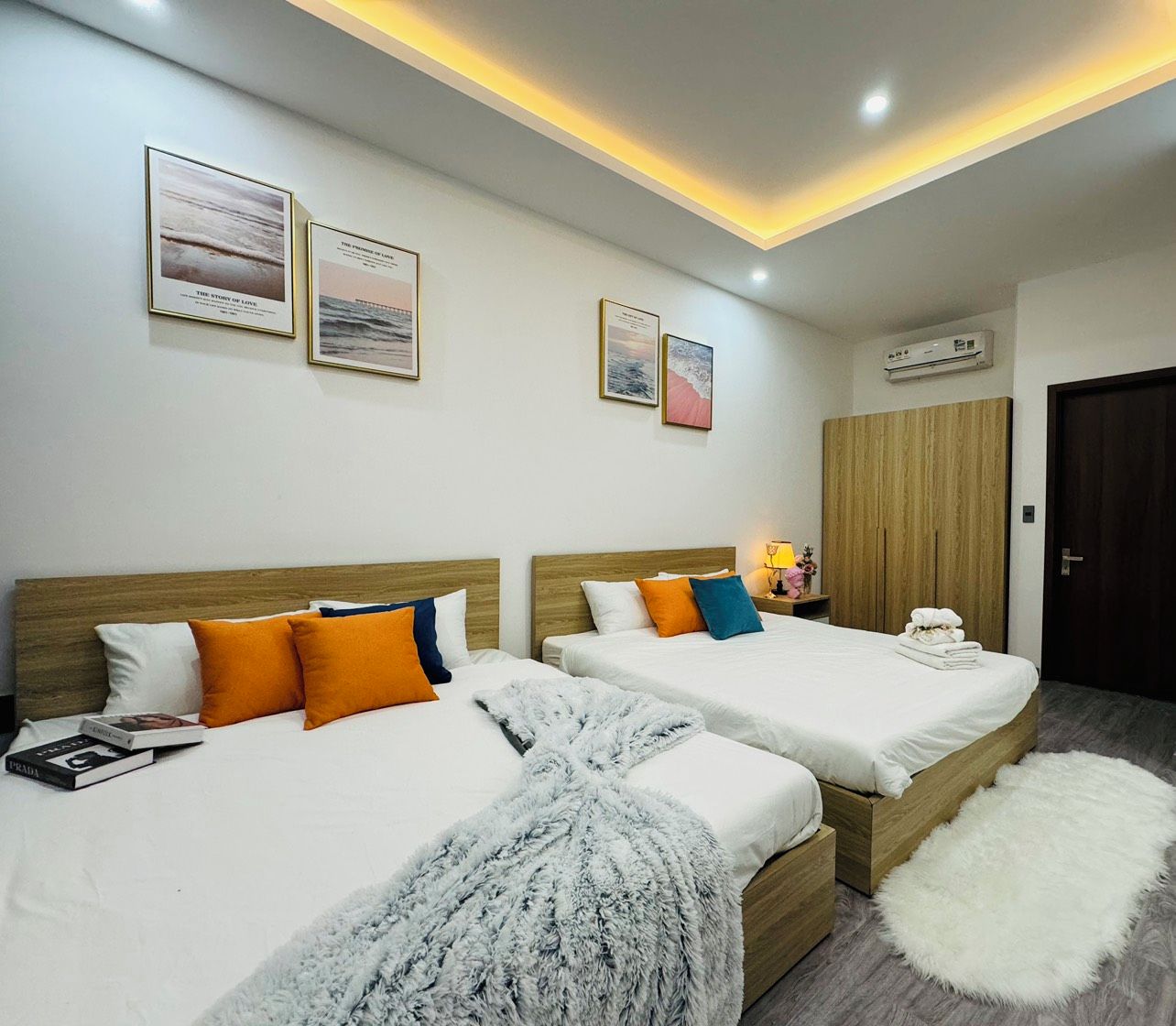 A hotel room with two beds, a wardrobe, a bedside table with a lamp, and framed pictures on the wall.