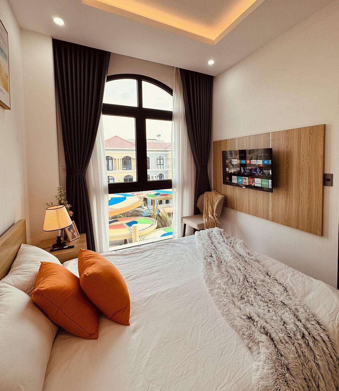 A hotel room with a bed, a nightstand, a lamp, a chair, a window with curtains, and a wall-mounted TV.