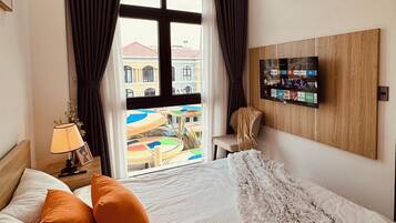 A hotel room with a bed, a nightstand, a lamp, a chair, a window with curtains, and a wall-mounted TV.