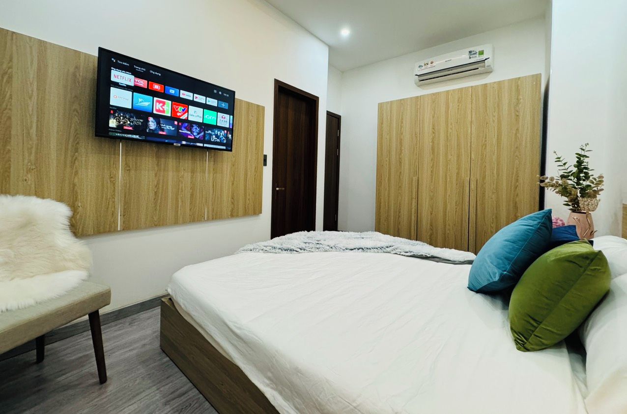 A hotel room with a bed, a mounted TV displaying various channels, a wooden wardrobe, and an air conditioning unit.
