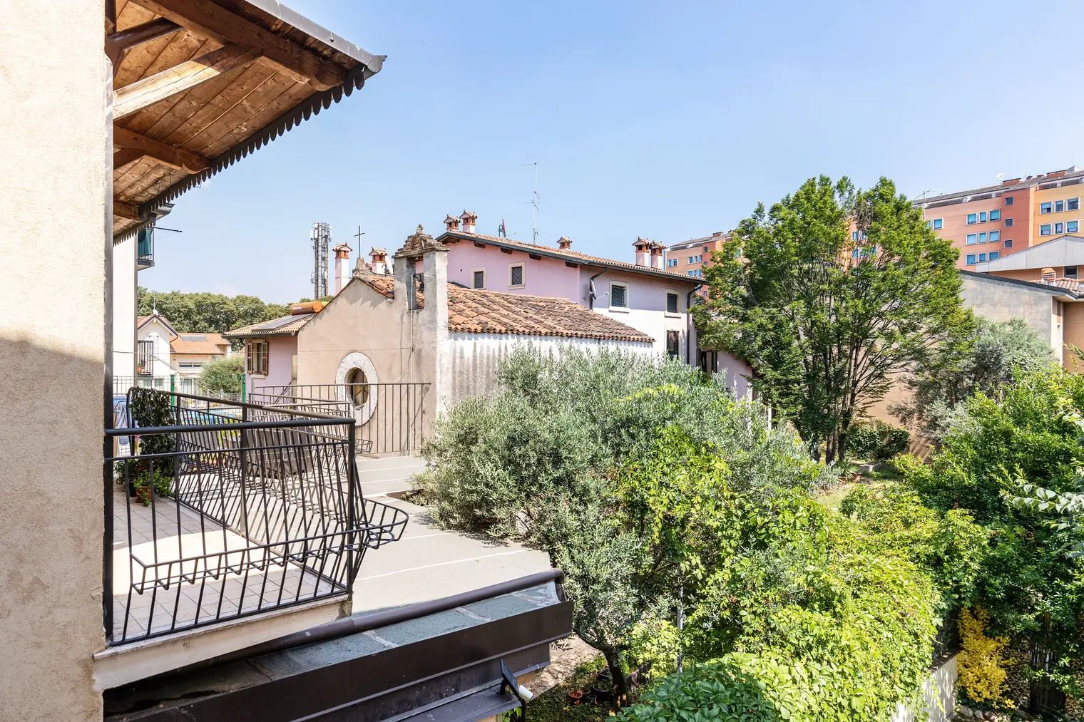 A residential balcony with a metal railing, a small garden area with various plants, and a view of neighboring houses with distinct architectural styles.