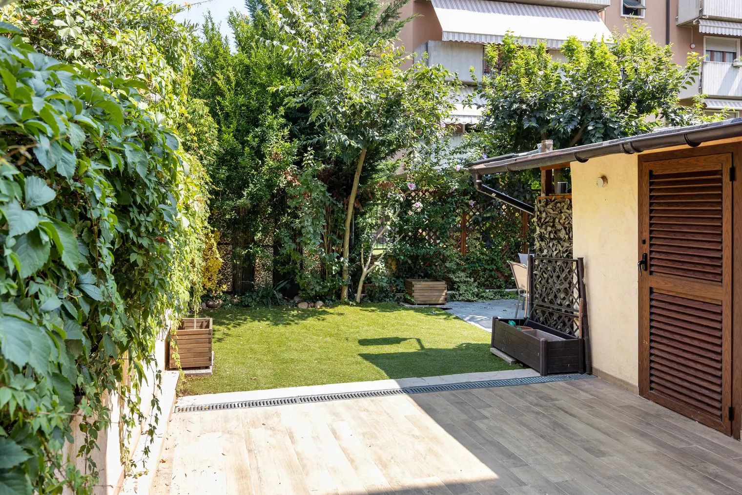 Un jardín residencial con una terraza de madera, un pequeño césped y un edificio con puerta y contraventanas de madera.