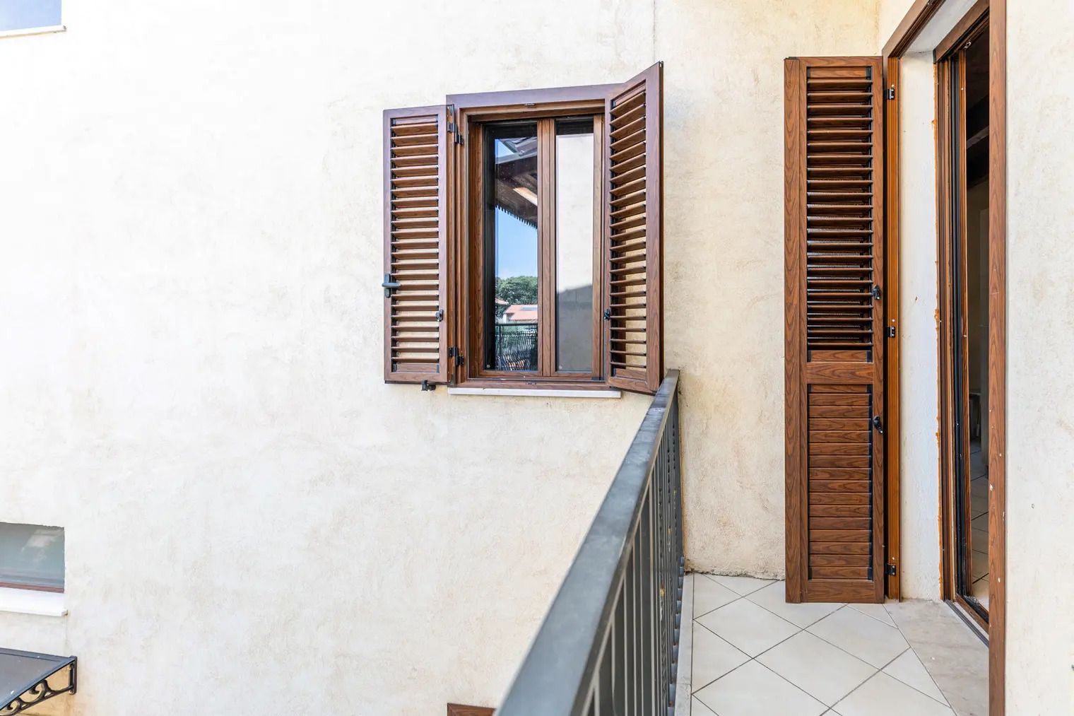 Un balcón con contraventanas de madera, suelo de baldosas y vista a un edificio al fondo.