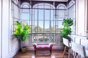 Une pièce avec un banc, deux plantes en pot, une table avec une chaise et une vue dégagée sur l’extérieur grâce à de grandes fenêtres.