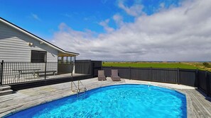 Een zwembad met een houten terras, een overdekte patio en uitzicht op een groen veld.