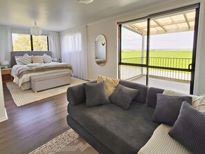 A bedroom with a bed, a sofa, a large window with a view, and a ceiling fan. - Pukehina Retreat: 
Private Pool, Pastures & Beach. (Pukehina)