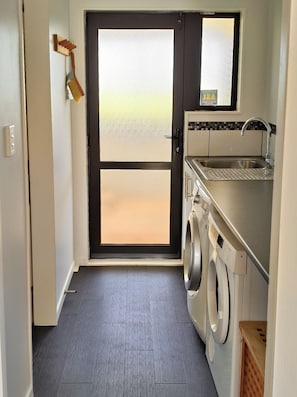 A laundry room with a washing machine, a sink, and a door leading to a balcony. - Pukehina Retreat: 
Private Pool, Pastures & Beach. (Pukehina)