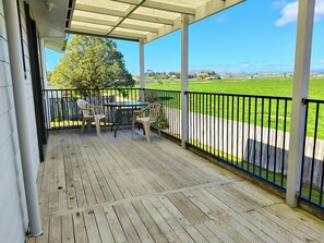 A covered outdoor area with a wooden deck, a table with chairs, and a view of a green field. - Pukehina Retreat: 
Private Pool, Pastures & Beach. (Pukehina)