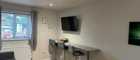 A modern kitchen with a dining area, a TV mounted on the wall, and a window with curtains.