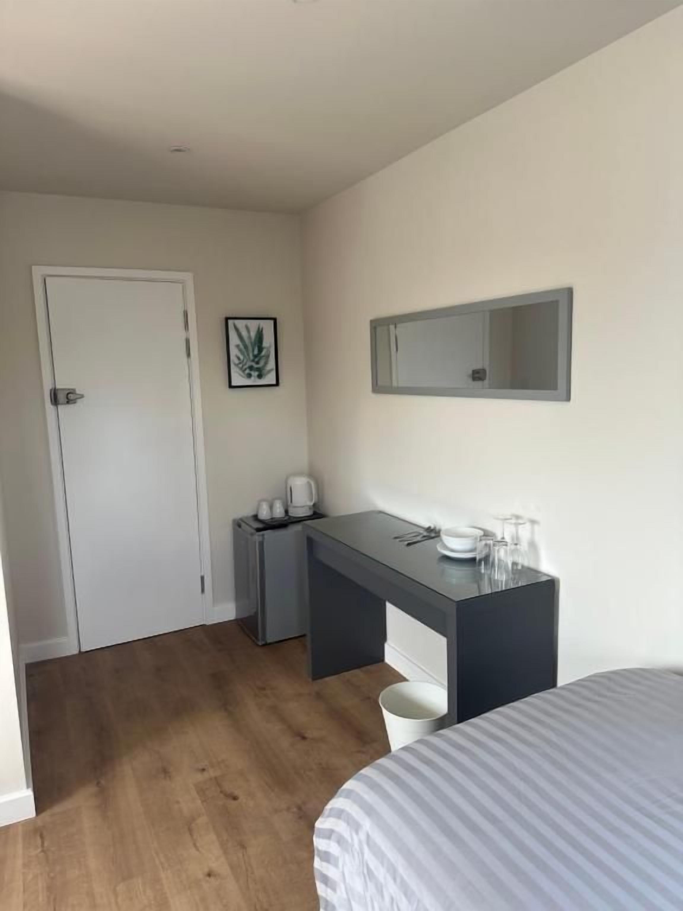 A small, clean room with a bed, a desk, a chair, a coffee maker, and a trash bin.