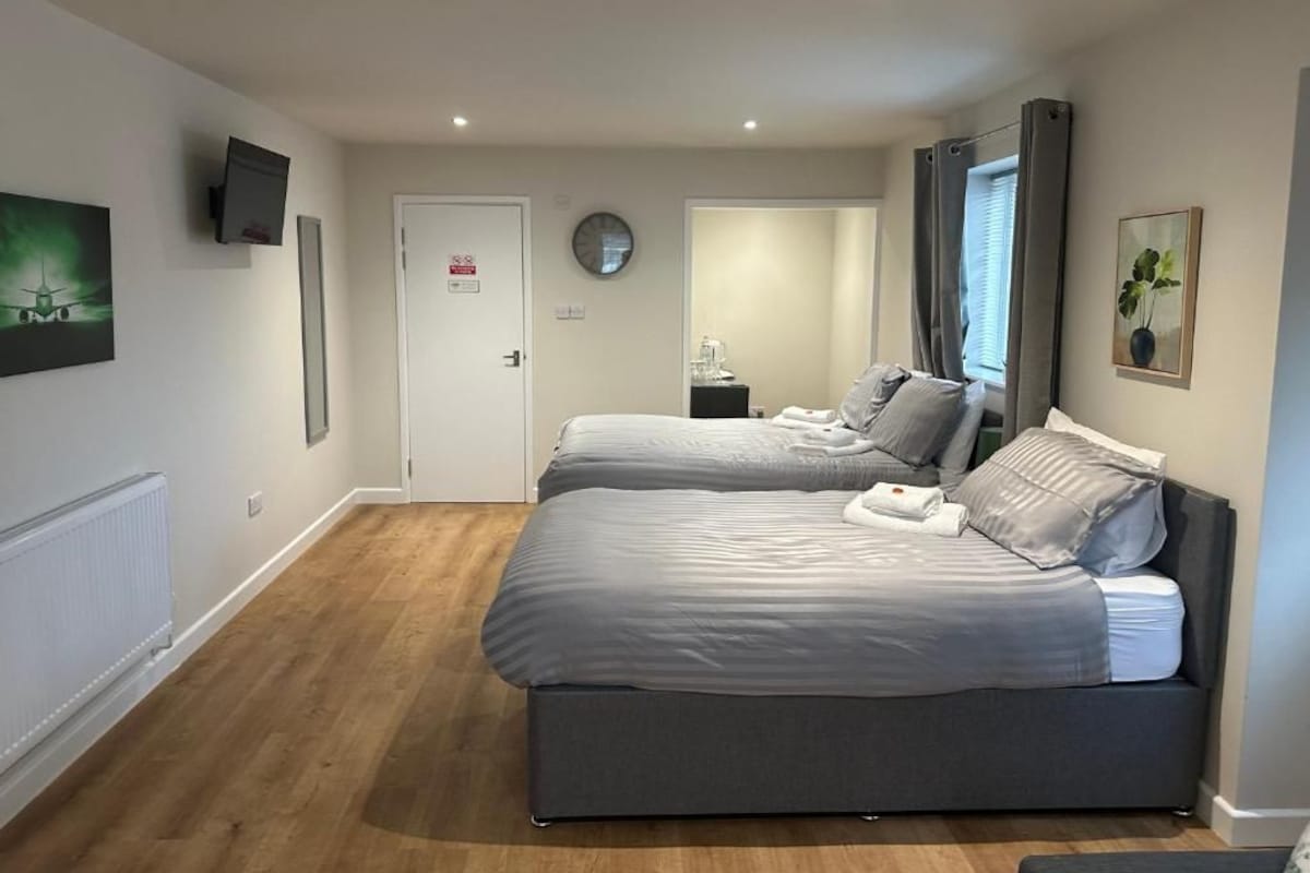 A hotel room with two beds, a bedside table, a clock, a radiator, and a television on the wall.