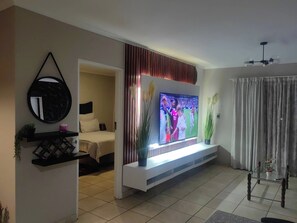 Un salon avec un téléviseur à écran plat fixé sur une console blanche, un lit avec une tête de lit, un miroir et une plante en pot.