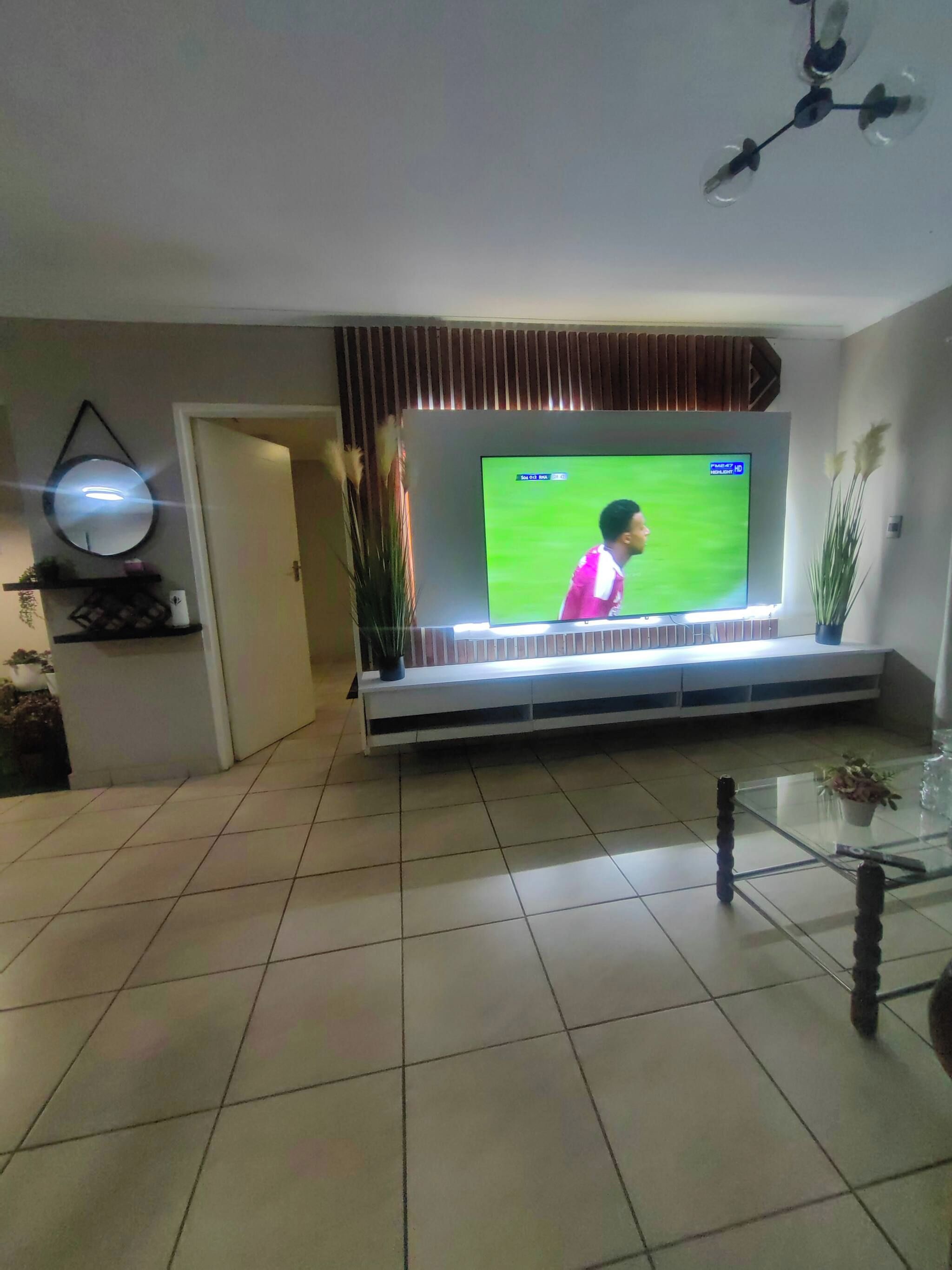 A living room with a flat-screen TV mounted on a wall, a glass coffee table, and a door leading to another room.