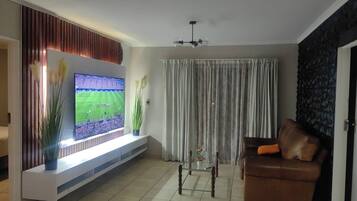 A living room with a flat-screen TV mounted on a wall, a brown leather sofa, and a large window with curtains.