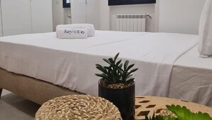 Een moderne slaapkamer met een bed, een nachtkastje met een bloempot, een geweven poef en een raam met uitzicht.