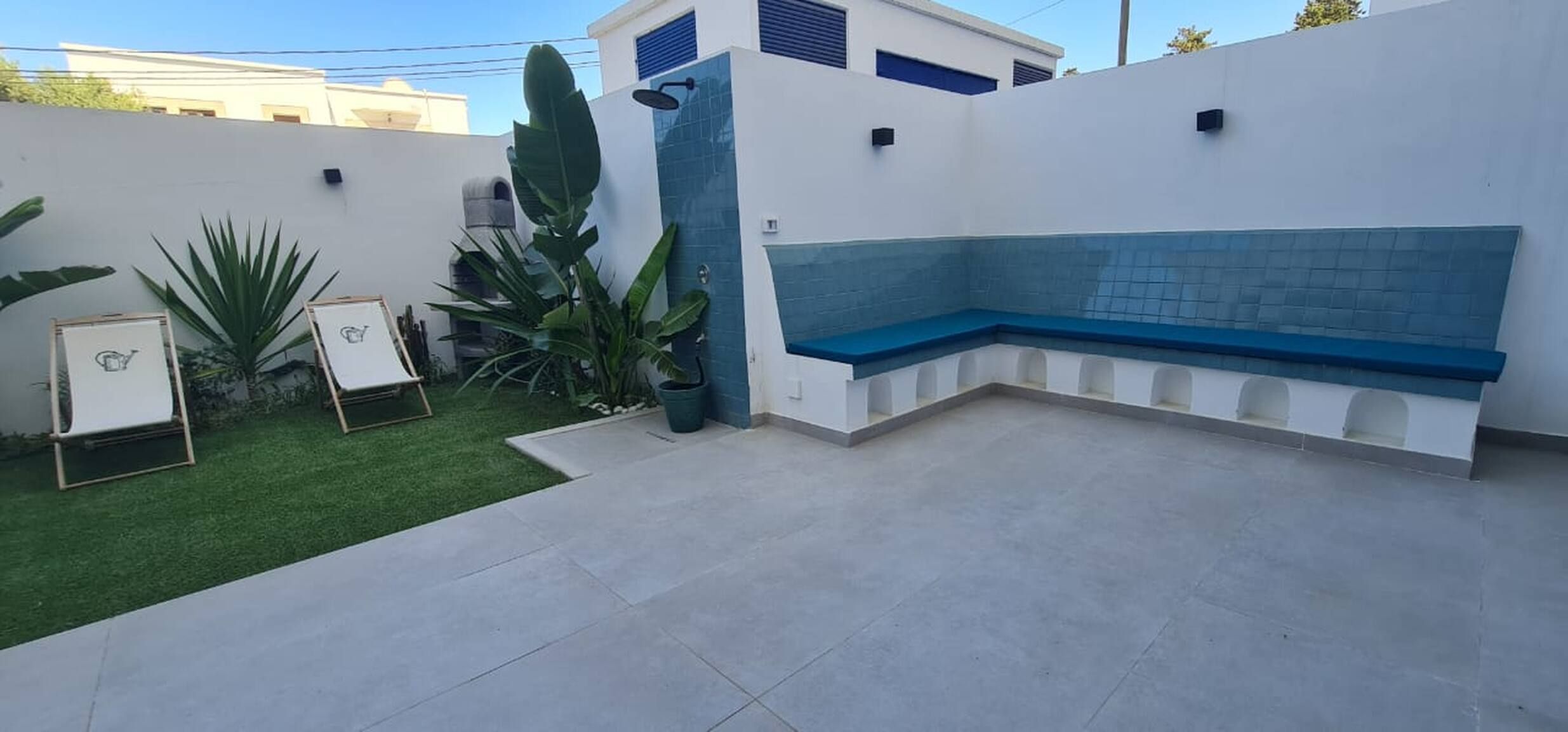 Un espace patio avec un banc bleu, un petit jardin avec des plantes vertes et un mur blanc avec une fenêtre.