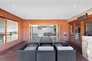 A modern outdoor patio with a glass-top table surrounded by wicker chairs, a brick wall, and a view into a living area. - Stylish Taylors Hill Kingdom (Taylors Hill)