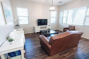 A modern living room with a leather sofa, a glass coffee table, and a flat-screen TV on a white cabinet. - Casa Encanto  - EV Charger, Pool, AC, H20 Filter (Goodyear)
