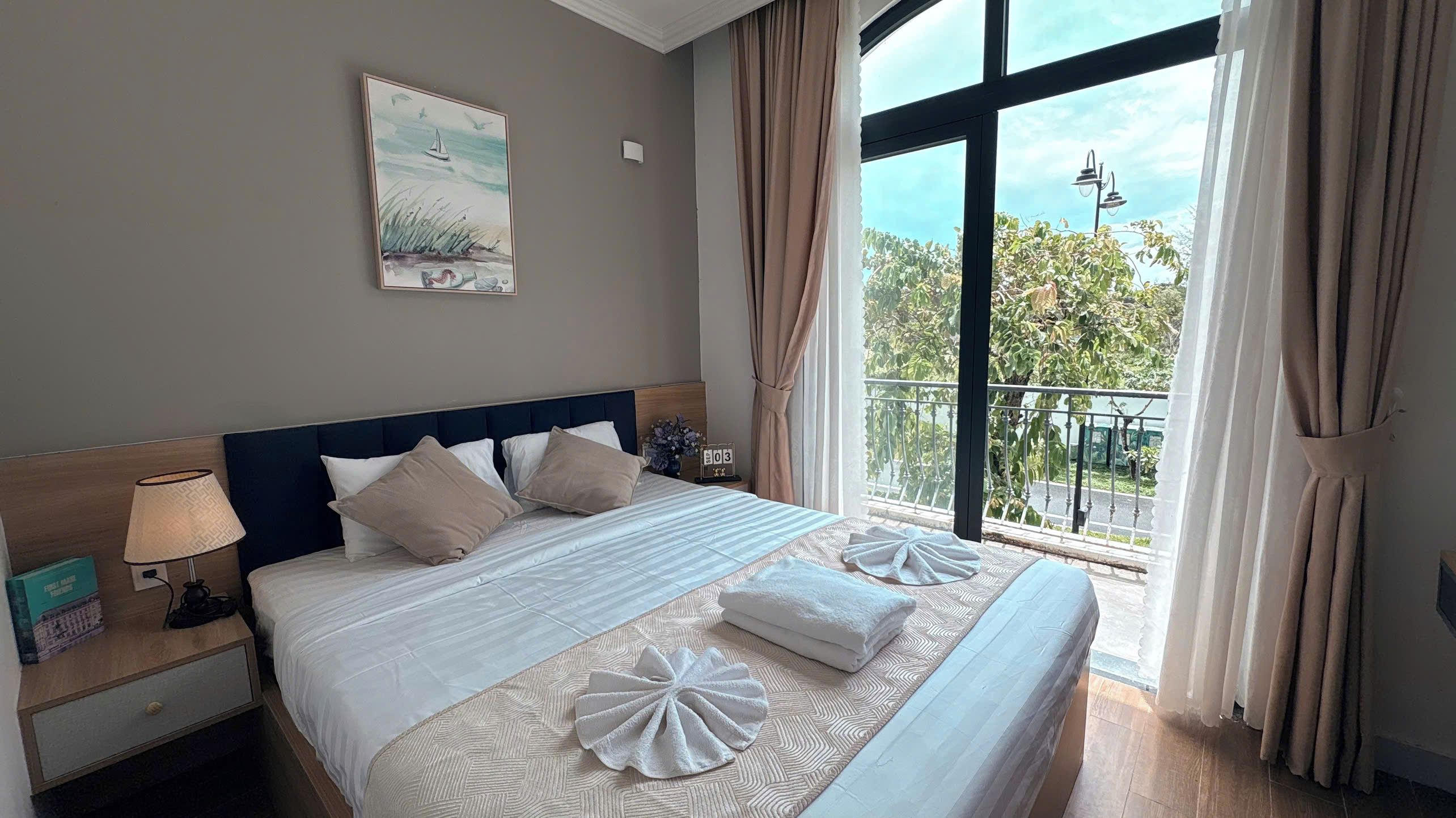 A hotel room with a large bed, a nightstand with a lamp, a painting on the wall, and a view of the outdoors through the window.