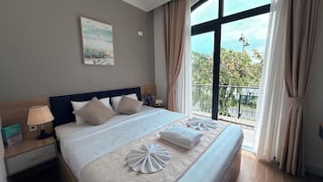 A hotel room with a large bed, a nightstand with a lamp, a painting on the wall, and a view of the outdoors through the window.