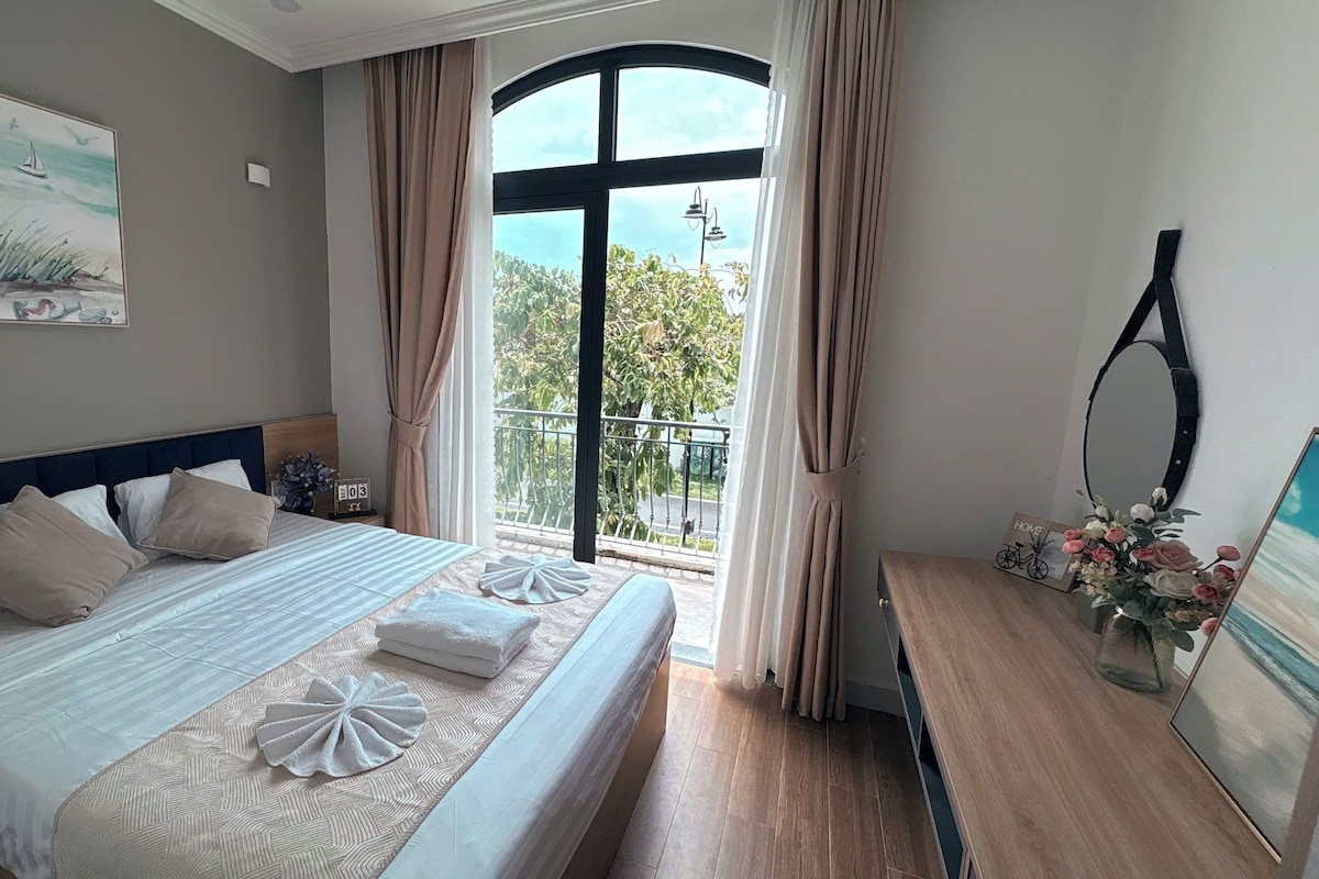 Habitación de hotel con una cama grande, vistas al exterior y una mesita con un jarrón de flores.