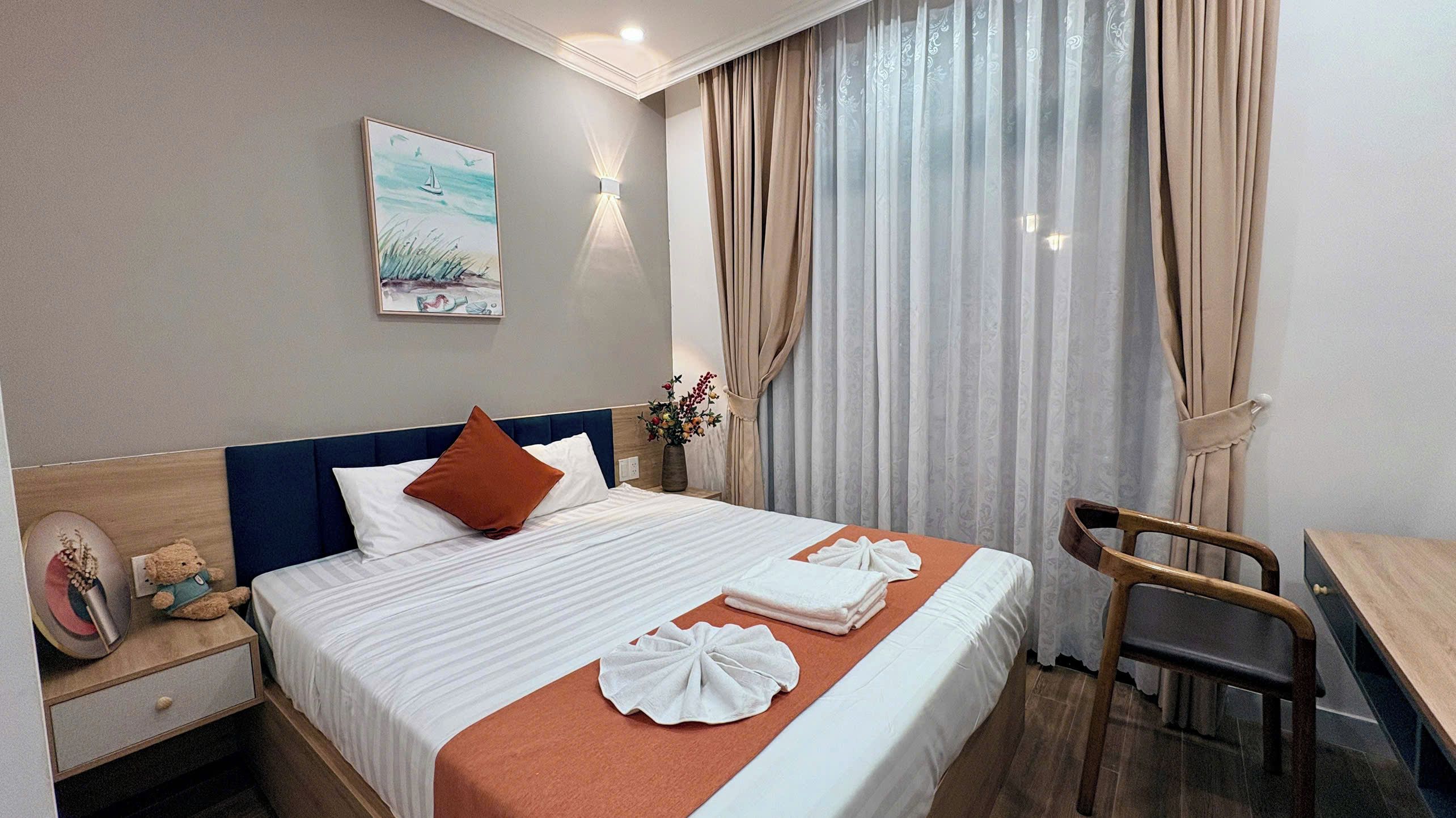 A hotel room with a bed, a desk, a chair, a nightstand, a vase with flowers, and a painting on the wall.