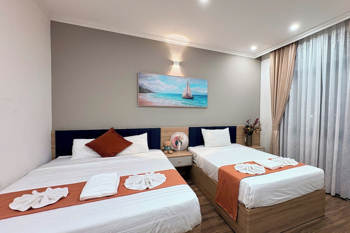 Una habitación de hotel con dos camas, un cuadro de un velero, una mesita de noche con un florero y un ventanal con cortinas.