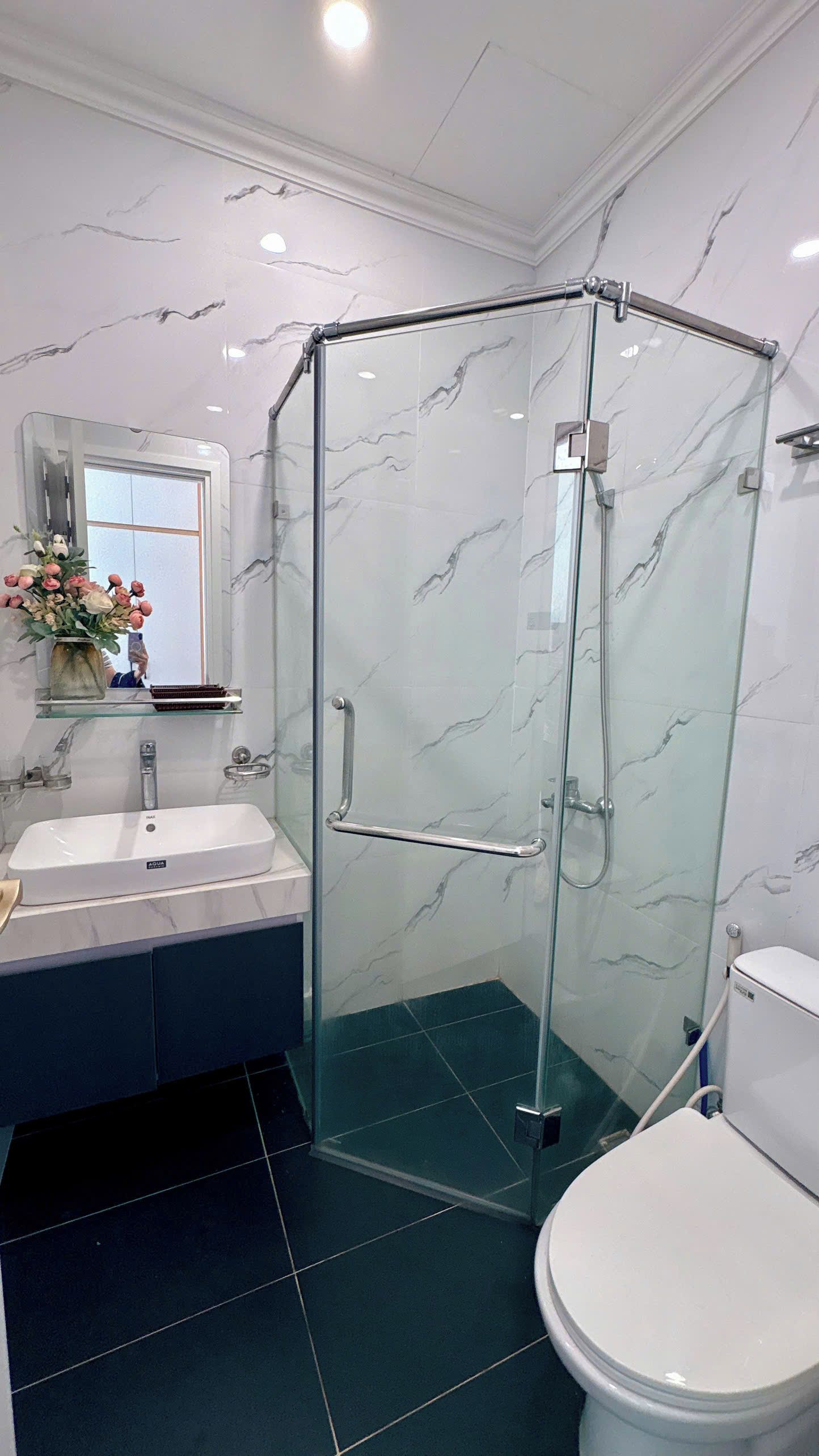 A bathroom with a glass-enclosed shower, a sink, a toilet, and a mirror with a vase of flowers.