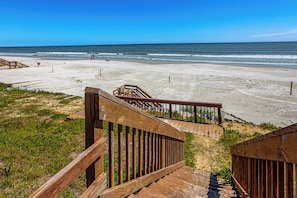 Eine Holztreppe führt zu einem Sandstrand, im Hintergrund das Meer.
