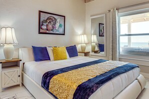A hotel room with a large bed, two bedside tables, a nightstand, a window with curtains, and a wall-mounted mirror. - Direct Daytona Beach Access & Views! Spacious Home (Daytona Beach)