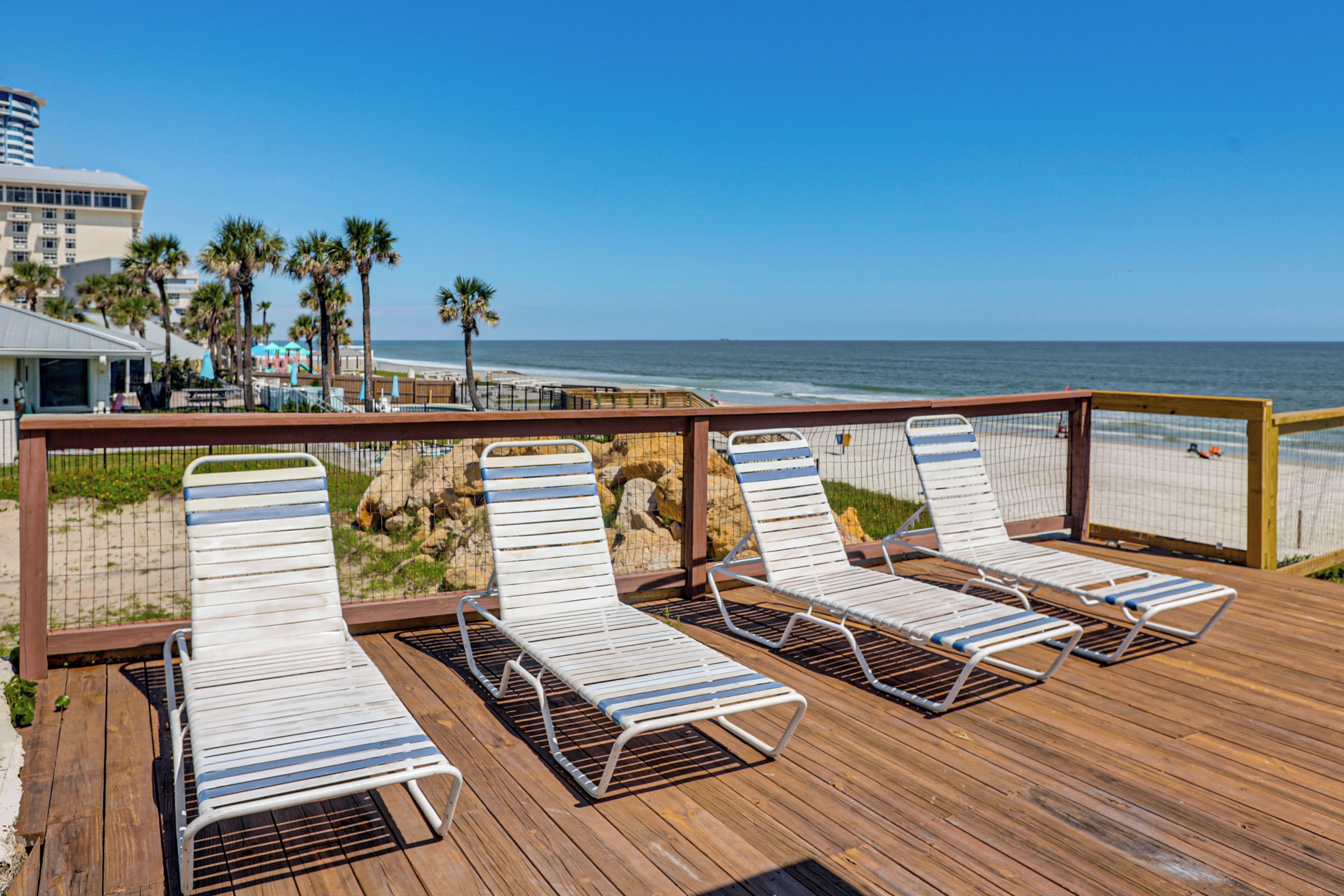 Une terrasse en bois avec des chaises longues donnant sur l’océan, une barrière rocheuse et une vue dégagée sur la plage et l’océan.