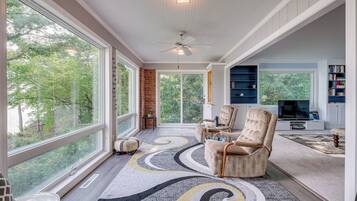 A spacious living room with large windows, a fireplace, a ceiling fan, and a television.