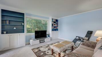 A living room with a television, bookshelves, a sofa, and a window with a view of trees.