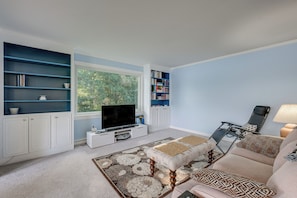 A living room with a television, bookshelves, a sofa, and a window with a view of trees.