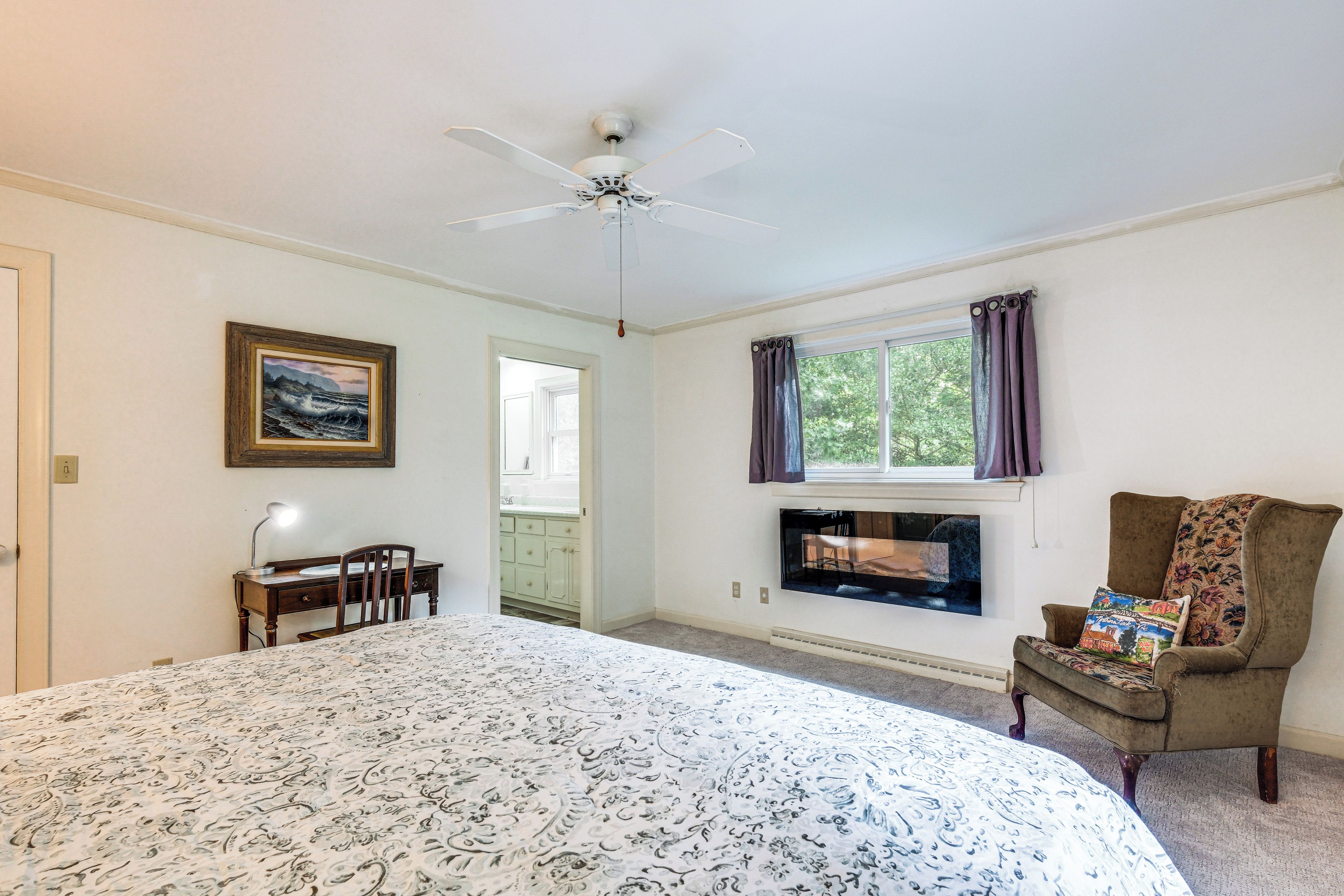 Une chambre à coucher comprenant un lit, un bureau, une chaise, un ventilateur de plafond, une fenêtre avec des rideaux et un tableau accroché au mur.