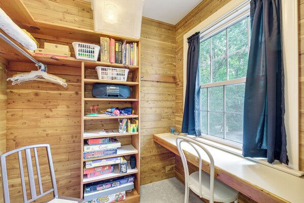A room with a wooden wall, a window with curtains, a desk, and a bookshelf with various items.
