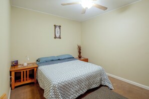 A bedroom with a bed, a nightstand, a ceiling fan, a wall-mounted picture, and a small vase with dried plants.