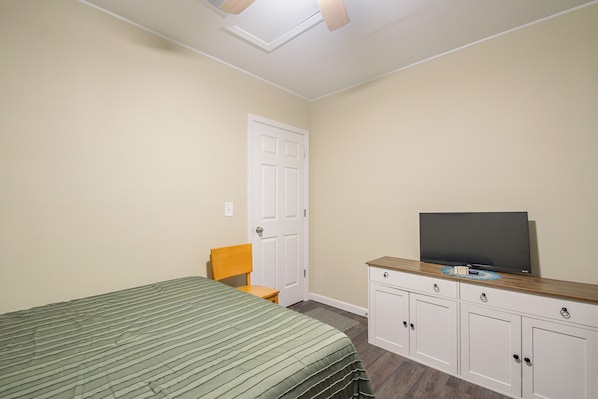 Un dormitorio con una cama, una silla, un televisor sobre un mueble y una puerta.