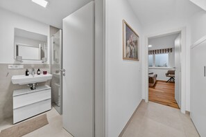 A modern bathroom with a sink, mirror, and storage, a hallway leading to a bedroom with a bed and window, and a painting on the wall. - Beachfront accommodation for 4 people in a new building with a terrace and private sauna - (Hörnum (Sylt))