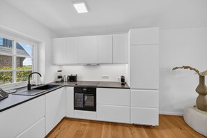 A modern kitchen with white cabinetry, a black dishwasher, and a window with a view of greenery. - Beachfront accommodation for 4 people in a new building with a terrace and private sauna - (Hörnum (Sylt))