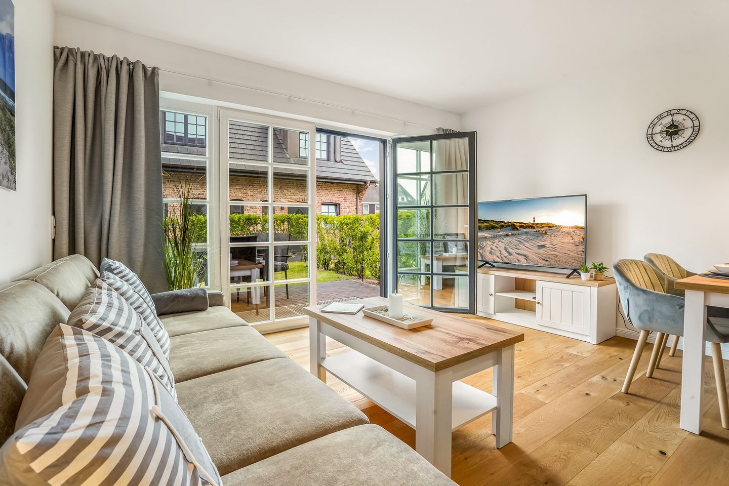 A modern living room with a sofa, a coffee table, and a TV. There is a balcony with a view of a brick building and greenery.